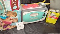 Easy Bake Oven displayed with its box and some accessories on top, including pink molds and a small aluminum pan, next to a box of GE soft white 100 watt bulbs.