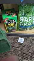 Miracle-Gro Lawn Food box, Scotts Grass Seed bag, and the manual spreader base visible on carpeted floor