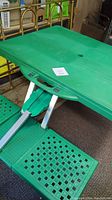 Green folding camping table unfolded showing umbrella hole and portion of attached plastic seats with perforations. Metal supports visible under table surface.