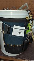 Front view of white bucket with dark blue fabric tool organizer attached. Tools and accessories including metal square ruler, saws, and cords visible.