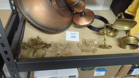 Photo of copper light fixture, glass taper holders, brass taper holder, and other brass accessories arranged on a shelf.