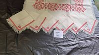 Full view of white linen tablecloth laid out with 6 matching napkins arranged. Red geometric embroidery on white fabric with fringe edges visible.