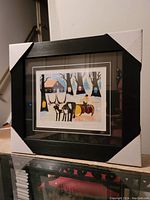 Framed Maud Lewis print in black wooden frame with corner protectors, showing full artwork and matting.