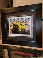Photo showing the entire framed print with the black wooden frame, double matting, and the colorful artwork inside depicting three black cats.