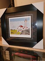 Framed Maud Lewis fine art print titled Lighthouse and Ferry shown with protective corner covers, displaying the full artwork, matting, and black wood frame.