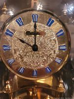Close-up of clock face showing ornate gold floral engravings and blue Roman numeral hour markers.