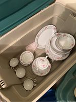 Photo showing stacked pink floral and silver-trimmed china including cups, saucers, plates inside plastic container