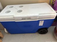 Blue and white wheeled Coleman cooler with lid featuring four cup holders. Black wheels and white pull handle visible. Cooler shows some dirt and wear indicating it needs cleaning.