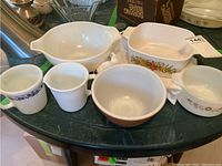 Top view of assorted CorningWare and Pyrex items including bowls, baking dish, milk glass cup and creamer on green table.