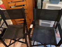 Two black folding director chairs side-by-side, showing full chair bodies including seats, arms, and backs made of mesh fabric with black metal frames.