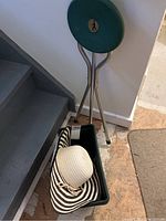 Wide-brimmed black and white striped sun hat in green plastic planter box with folding seat behind it on basement stairs.