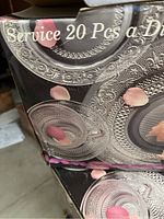 Box close-up showing embossed patterned glass plates and cups with rose petals for decorative effect. Displays 'Service 20 Pcs a Dinner' text.