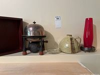Overview shot showing all items in the lot lined up on a shelf: wooden tray, cast iron fondue warmer with silver plate dome, clear glass dish, speckled pottery jug, red glass vase with silver base.