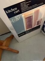 Box of the kitchen cart showing features and materials such as maple finish, solid wood cutting surface, metal castors, and wire baskets.