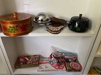 Wide view of all items on shelves showing wooden box, metal covered dish, fondue pot, wooden utensils, cookie cutters, swizzle sticks, coaster, and hostess mold box.