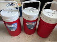 Three red Coleman thermos containers with white lids and a Little Playmate cooler seen in background.