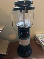 Full view of propane lantern showing black metal top, glass enclosure, handle, base with control knob and instructional label