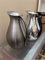 Two aluminum jugs side by side showing contrast in finish and handle material.