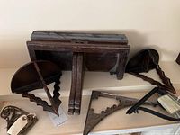 Three wooden wall sconce shelves and four metal shelf brackets on a white shelf. Two dark metal brackets have a simple carved design; two brass brackets with ornate scroll pattern.