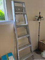 Side view of the aluminum step ladder leaning against wall near window, showing four wide steps and top tray platform with round holes.