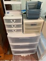 Stacked plastic storage drawer units with one having three large drawers, another with three smaller drawers, and a stack of six gray plastic baskets on top.