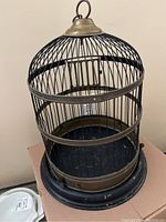 Front-side view of vintage metal birdcage showing cylindrical wire cage, brass top with hanging loop, and removable base.