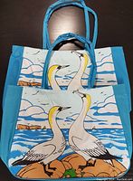 Two canvas beach bags with turquoise sides and handles; front panels depict two gannets by the sea with fish, rocks, waves and sky.