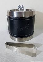 Front view of the Bacardi ice bucket covered in black leather with embossed Bacardi text and the stainless steel top rim and base with ice tongs in front.