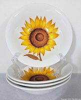 Four white ironstone plates with a detailed yellow sunflower pattern with brown center and leaf, displayed stacked with one standing upright.
