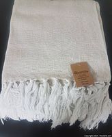 Folded beige and white cotton throw with fringe, showing fabric texture and brand tag.
