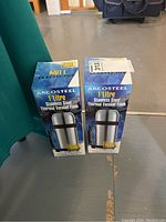 Two boxed Arcosteel 1 litre stainless steel thermal vacuum flasks placed side by side on floor with green chair in background.