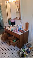 Wide view of bathroom vanity with countertop showing cleaning supplies, soaps, spray bottles, wooden paper towel holder, plastic storage containers, and open vanity drawers showing additional supplies.