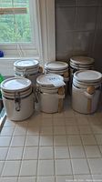 Six white ceramic canisters with airtight locking lids on a counter by a window, each with a small wooden spoon attached. All appear clean but used.
