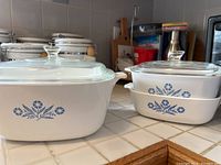 Set of 3 white Corningware dishes with blue cornflower pattern, 2 smaller dishes stacked with lid, and one larger dish on the left with lid.