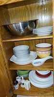 Photo of stainless steel mixing bowls stacked along with white Corelle dishes and assorted ceramic bowls, spoons, and shakers inside a wooden cabinet.