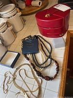 Photo showing four necklaces including faux pearl strands (white and black), a wood beaded necklace, blue jewelry box insert and a glossy red heart-shaped jewelry box.