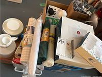 Box containing kitchen linens, rolls of mesh or fabric, and small kitchen accessories.