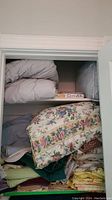 View of upper closet shelf with folded light gray blankets, floral pillow covers, and stacks of folded sheets in various colors such as yellow and green.