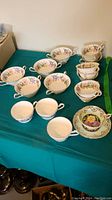 Overview of all cups and saucers including double-handled cups, Country Lane cups, and ribbed cups laid out on a green table covering.