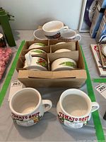 Top view showing several white ceramic soup cups with handles inside a cardboard box with dividers.