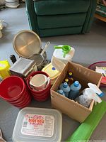 Photo showing a stack of red plastic bowls, metal pots and pans, plastic containers, cleaning liquids in a box, and a margarine container for storage.