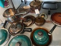 Overview of brown amber Visionware glass cookware with black handles and some green enamel pots. Includes lids on almost all cookware pieces.