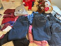 Photo showing multiple clothing items including folded jeans, shirts, pyjamas, flannel nighty, and housecoat on a bed along with several teddy bears in background.