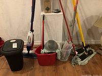 Cleaning supplies including mops and buckets arranged against a wall showing bucket with wringer, mesh waste baskets, two flat mops and one string mop, and black plastic trash basket with lid