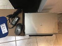 Photo showing HP EliteBook laptop closed, black Verbatim wireless mouse, HP power adapter with cable, all on a cardboard box next to a small white step stool.