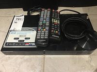 Top view of Samsung BD-F6900 DVD/Blu-ray player with two remotes and cables on tiled floor.