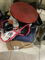 Close-up photo of assorted exercise equipment including balance disc, skipping rope, and resistance bands piled on a box in a basement setting.