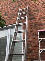 Aluminum extension ladder leaning against a brick wall outdoors, extended and showing multiple rungs and locking mechanisms.