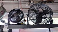 Two fans on a shelf: one large black metal Honeywell fan about 16" in diameter, one smaller black plastic fan.