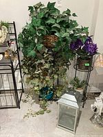 Overview photo showing the faux plants, metal lantern, multiple plant stands, and other smaller plant pots grouped on floor against a white wall.